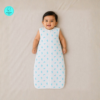 A smiling South Asian baby lies on a soft beige blanket wearing a lightweight white muslin sleeping bag with a front zipper and a small blue whale. The sleeveless sleeping bag has a slim, straight silhouette with a rounded bottom, while the baby's arms rest comfortably at the sides in a bright, softly lit overhead photograph.