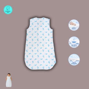 A smiling South Asian baby lies on a soft beige blanket wearing a lightweight white muslin sleeping bag with a front zipper and a small blue whale. The sleeveless sleeping bag has a slim, straight silhouette with a rounded bottom, while the baby's arms rest comfortably at the sides in a bright, softly lit overhead photograph.